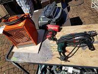 Lot showing orange portable heater, Milwaukee red heat gun, and green Metabo hammer drill on plywood board outdoors under daylight.