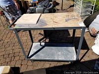 Full view of metal utility cart on paved surface showing wood top and lower metal shelf with wheels.