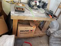 Top and front view of the workbench showing metal top with scratches and holes, wood base, and plywood bottom shelf. Cardboard boxes stored underneath the bench.