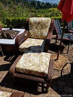 View of wicker chair and ottoman with leaf patterned cushions placed on a patio with garden background