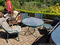 Outdoor round glass top patio table surrounded by four patio chairs with cushions, situated on a paved patio.