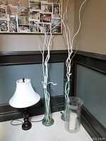 Photo of the lamp, two tall green glass vases with white decorative branches, and a clear glass hurricane candle holder with white pillar candle all arranged together on a hardwood floor near a wall.