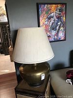Full view of large brass lamp showing bulbous polished brass base with cream fabric lampshade and surrounding room context