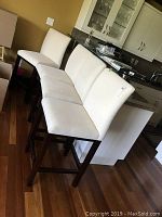 Four upholstered counter height chairs lined up in kitchen area showing front and side views of the set, cream fabric upholstery and dark brown wooden legs.