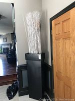 Full view of tall black metal decorative vase with white decorative sticks placed near a wooden door and wall corner.