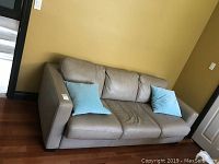 Full view of the light grey leather sofa bed with two blue throw pillows on a wooden floor against a yellow wall.