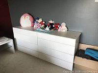 Front view of white 8-drawer dresser with plush toys on top, showing full width and drawer fronts.