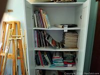 White cabinet with shelves containing books, magazines, Beware Of Dog signs, airbrush, and compressor.