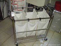 View of the chrome frame laundry hamper with three white removable bins inside and a lower wire shelf. The hamper is on four caster wheels and placed on tiled floor.