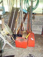 Photo showing various long-handled garden and outdoor tools including rakes, shovels, brooms, roof rake, axe, pickaxe, red fuel can, red metal toolbox, and hose reel stand with yellow extension cord.