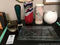 Wide view showing a rectangular clear glass tray with dragonfly design and five vases including a red glass vase, tall green glass vase, floral ceramic vase, white ceramic pitcher, and a small marbled glass vase.