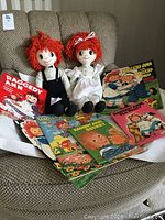 Two Raggedy Ann and Andy dolls seated on chair with multiple vintage Raggedy Ann and Andy books spread around them.