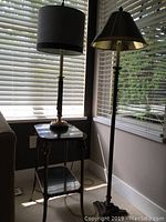 Side table, composite table lamp with black shade, and tall metal torch floor lamp near window with blinds.