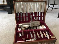 Opened wooden case showing silver plated cutlery including knives, forks, spoons, and serving pieces neatly arranged in red velvet interior.