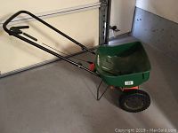 Full side view of the Scotts Turf Builder fertilizer spreader showing the green hopper, metal handle with adjustable lever, and single wheel.