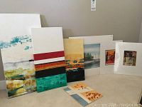 A group photo showing six unframed art pieces in varied sizes leaning against a gray wall, comprising two larger canvases with textured abstract designs and four smaller prints or paintings with diverse themes.