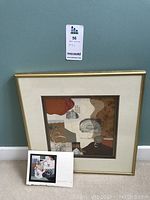Framed mixed media collage artwork in a gold frame with cream mat. The artwork is in earthy colors with abstract textured paper designs. Original artist's postcard with handwritten note is also shown.