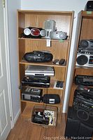 Full view of bookshelf containing a mix of boom boxes, VCRs, and a box of clock radios at bottom shelf.