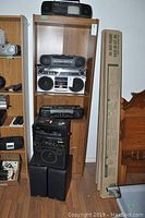 Shelf with various vintage boom boxes and stereo equipment including a small TV on top and separate speakers on floor