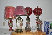 Complete view of all lamps and shade on a wooden surface, showing the variety of styles and colors including two tall lamps with red spherical glass shades and two vintage oil lamps along with ceramic lamps.