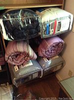 Six comforters stacked and stored on shelving, four sealed in plastic bags (two on top shelf and two on bottom) and two purple striped rolled comforters not in plastic on middle shelf.