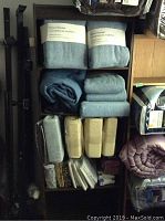 Shelf with multiple packaged bedding items including fleece blankets, folded blankets, pillowcases package, plastic-wrapped sheets, and a purple comforter.