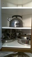 View of stainless steel kettle on upper shelf and pots, pans, lids on lower shelf inside white cabinet