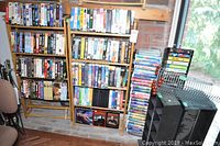 Photo showing two wooden shelves stocked with VHS tapes, a tall stack of loose VHS tapes in a clear plastic rack, and a few metal/plastic VHS organizers.