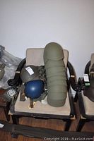 Photo showing nine green helmet liners stacked upright on a chair with three military style helmets placed around them, with canteens and military pouches visible to the side.