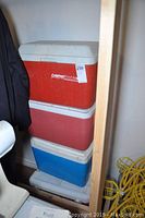 Stack of four coolers, two red and one blue, with white lids in a corner.