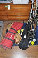 Two bat bags, one red Rawlings and one black, with multiple bats inside, several baseballs and softballs, and leather gloves placed outside the bags.
