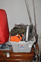 Photo of plastic tub filled with hunting clothes including camouflage and orange vests and other clothes, with fishing rods placed behind it.