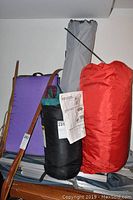 Camping gear including red sleeping bag sack, purple sleeping mat, folded camping chair, and tent poles bag with instructions.