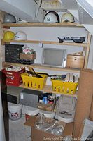 Storage room wood shelving showing safety helmets on top shelves, plastic organizer drawers with hardware items, bins containing painting supplies, shoe shine kit, and other containers on lower shelves.