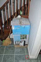 Front view of sealed Commercial Cool 12,000 BTU portable air conditioner box leaning against wall with broom and staircase behind.