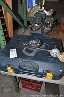 Photo showing the closed plastic carrying case with metal lance accessory on top and additional item partially visible in background.