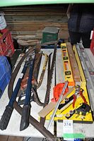 Photo showing an assortment of hand tools including levels, hand saw, crowbar, chain cutters, tape measure, angle square, and hand saw frame on a table with a wooden wall background.