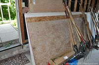 Stack of drywall sheets in a garage leaning against a wall near garden tools.
