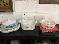 Lot of six kitchen dishes on black table against white brick wall, including Pyrex casseroles and clear glass bakeware dishes