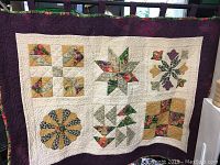 Front view of the full quilt showing six patchwork blocks with floral and geometric patterns, cream background, and purple border.