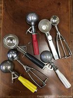 Top view of seven vintage metal ice cream scoops with different handle colors including red, yellow, black, and white, all laid out on a wooden table.