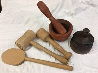 Five wooden kitchen utensil pieces comprising butter mold, mortar and pestle, meat tenderizer mallet, wooden pestle, and spatula on light colored fabric.