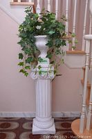 Full view of white fluted pedestal with faux ivy plant in urn planter on top, positioned against a staircase.