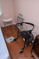 Black Nexus Walker with padded seat, hand brakes, metal basket under seat, positioned next to a folded lightweight walker and a heating blanket on the floor.
