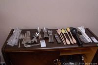 Wide view of multiple metal flatware pieces including forks, knives, spoons, tea spoons, and some plastic wrapped cutlery on a dark wood surface.