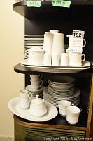 Photo of two shelves with various white fine china dishes including plates, cups, pitchers, and condiment containers.