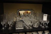 View showing 19 crystal sparkling wine flutes arranged in rows, with a crystal decanter and napkin holders in front, and salt and pepper shakers, small glasses and ice bucket behind, all on a shelf with a large silver tray in the background.