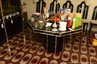 Black octagonal dining table with gold trim, top and base visible, surrounded by chairs and various kitchen items placed on top.