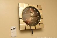 Front view of Bulova mid-century modern style square wall clock with metallic gold-tone casings and a brown gradient sunburst dial, showing the clock face and pendulum.
