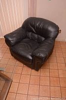 Front angled view of black leather armchair on tiled floor near vertical blinds showing padded arms, wide seat and back cushion.
